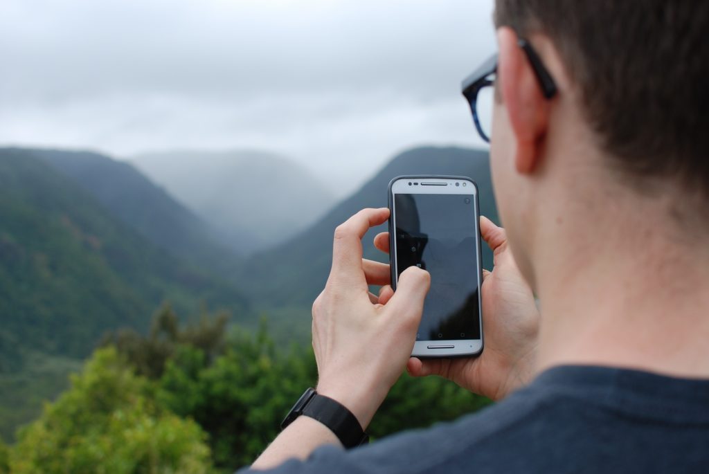 Das Foto zeigt einen jungen Mann, der mit seinem Smartphone die Landschaft fotografiert.